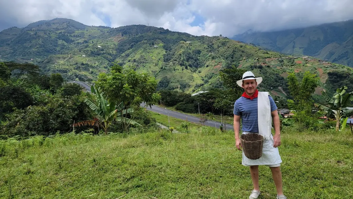 Jeffrey Booth smiling in a straw hat and red neckerchief, holding a woven basket on a green mountainside